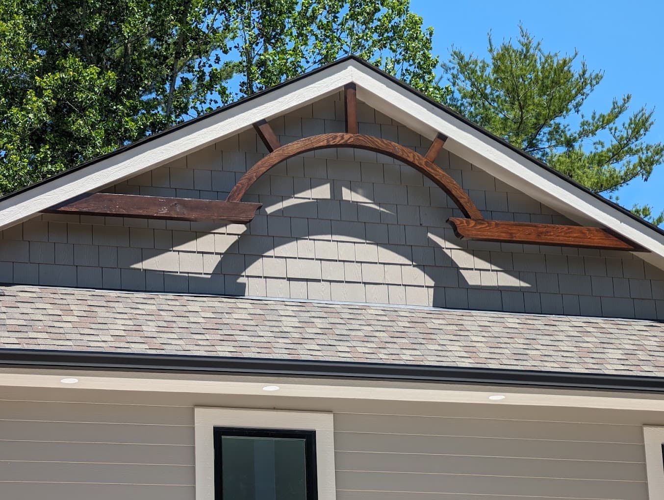 A single arched wood bracket, stained dark brown, spanning the peak of a gable end finished in gray shingle siding, with a short horizontal kicker return on each side.