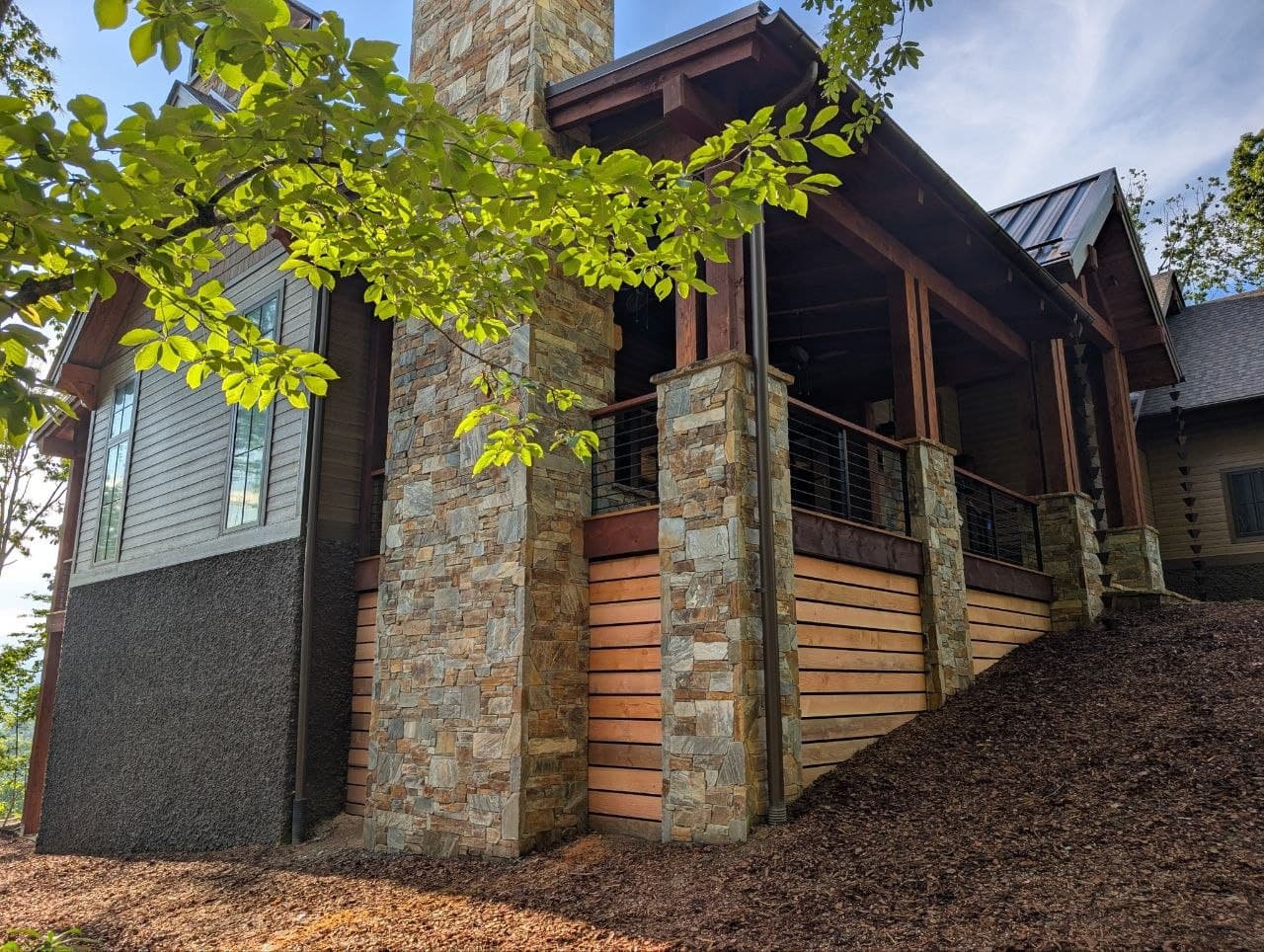 Horizontal cedar skirting run tight to stacked-stone columns beneath a covered porch, with cable railing above