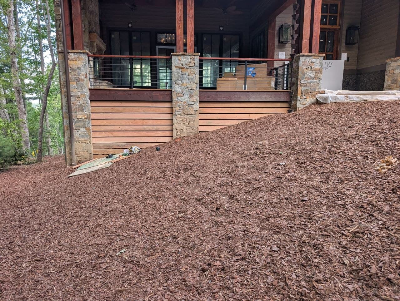 Stone porch columns with stained horizontal skirting boards run between them, cable-rail deck above, house still under construction with a mulched bed at grade.